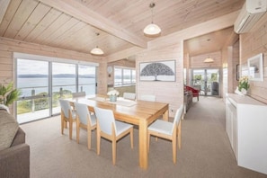 A modern interior with a wooden dining table, chairs, and a view of the sea. - Aotea Harbour Beach House (Kawhia)