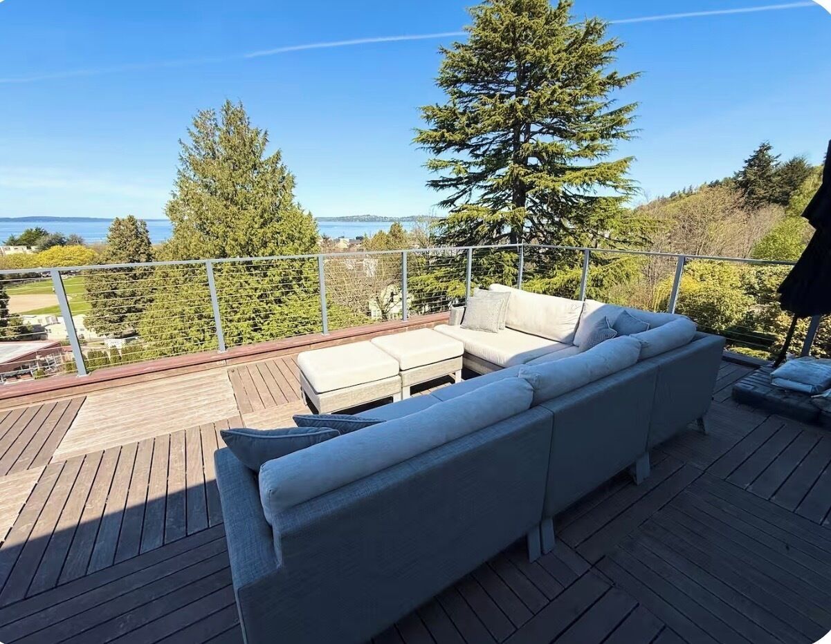 Une terrasse avec un canapé et un pouf, donnant sur une étendue d’eau et des arbres.