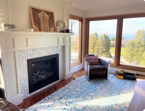 A room with a fireplace, a large window with a view, a patterned rug, and a leather chair. - Seattle Luxury VIEW House, Walk to Beach, Park, Forest, Tennis Court (Seattle)