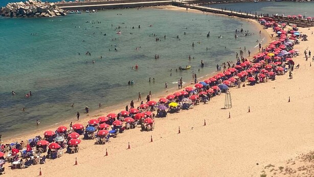 Una platja amb un pontó rocos i aigua blava cristal·lina, i nombrosos paraigües.