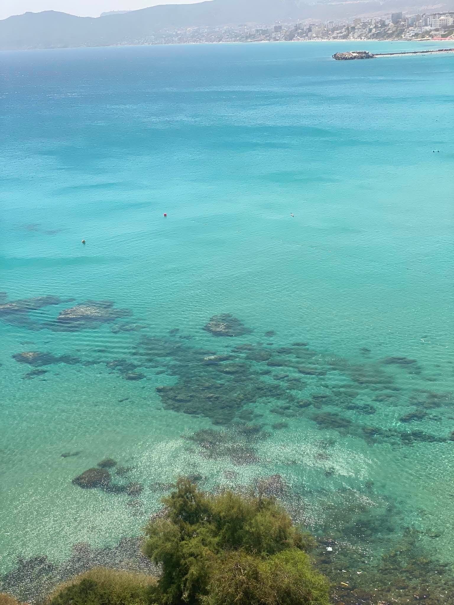 A coastal view with clear turquoise waters, rocky outcrops, and distant buildings.