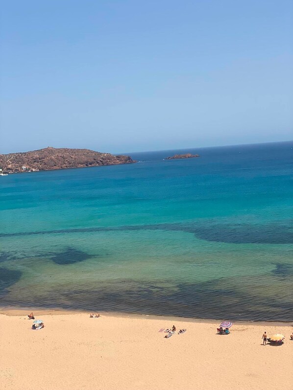 Una playa con agua turquesa cristalina, orilla arenosa y un afloramiento rocoso en la distancia.