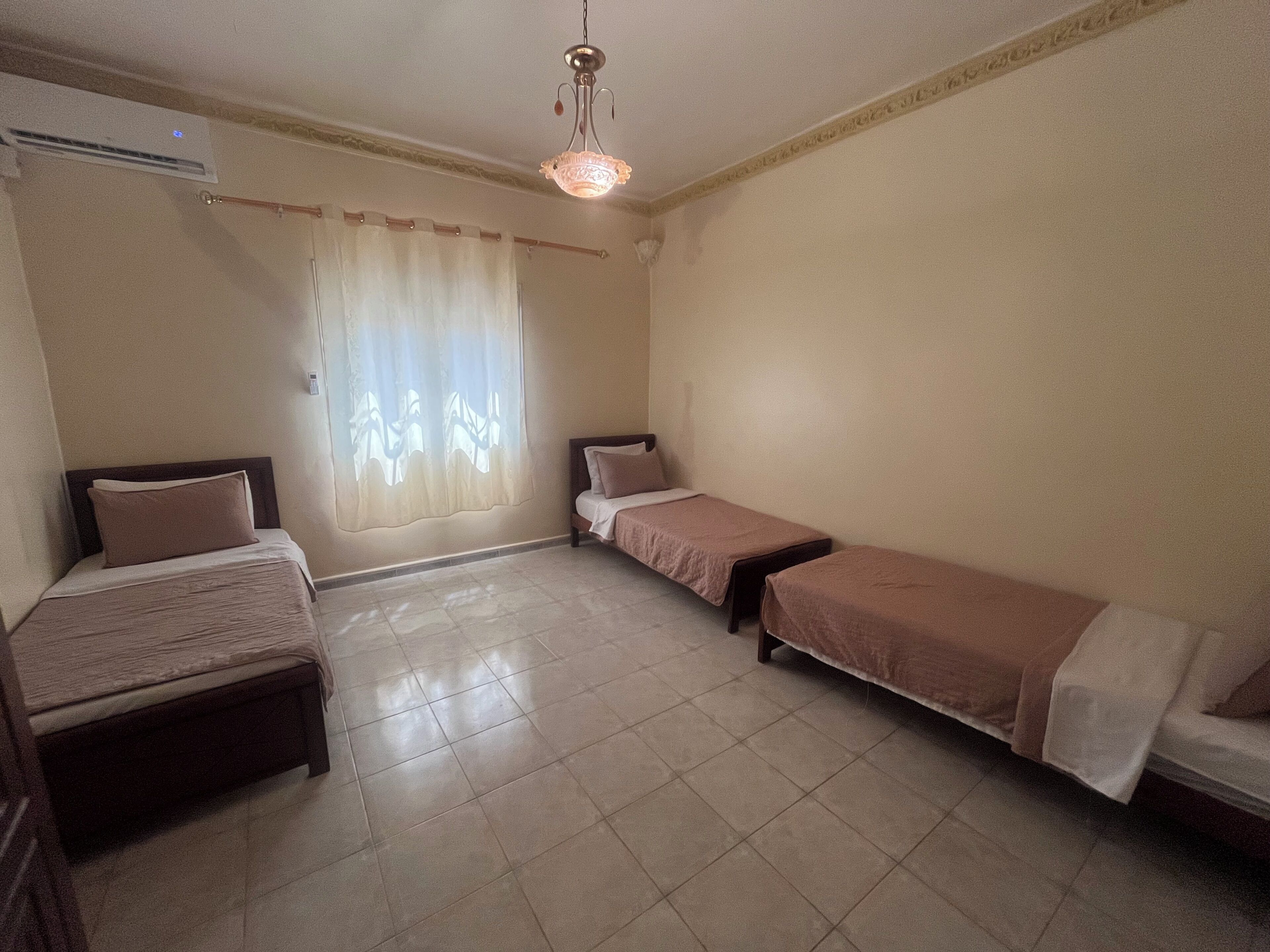 Een kamer met twee bedden, een raam met gordijnen, een plafondlamp en een airconditioning.