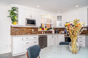 Una cocina moderna con armarios blancos, salpicadero de ladrillo y un comedor con mesa y sillas de vidrio.