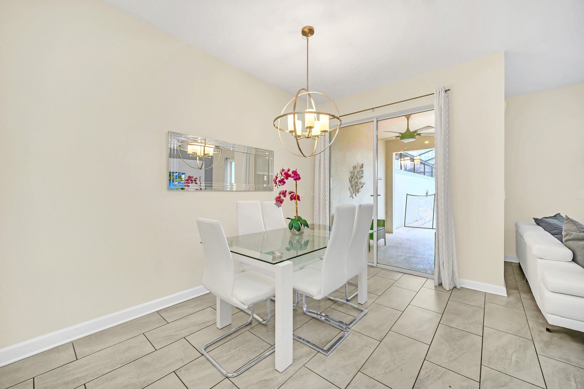 A dining area with a glass table, white chairs, a chandelier, and a view through a glass door to a balcony.
