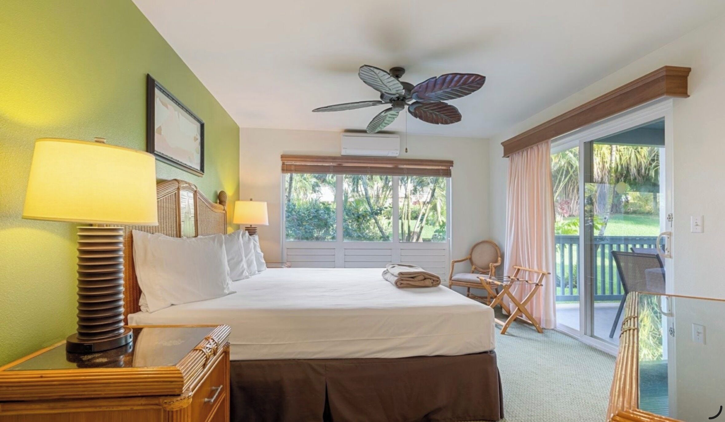 A hotel room with a bed, two bedside lamps, a ceiling fan, a window with blinds, a chair, and a balcony with a view of greenery.