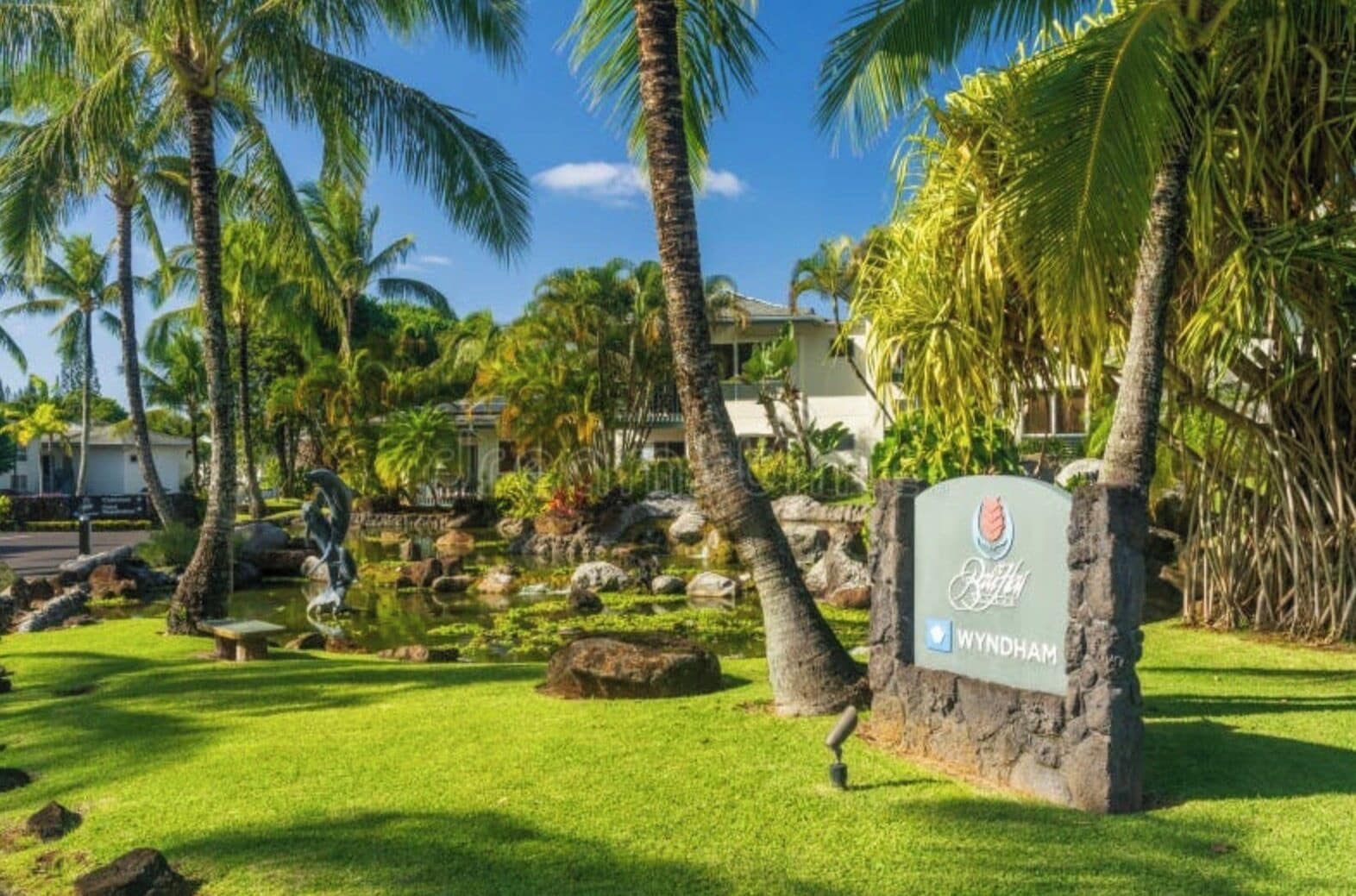 A tropical resort with a sign reading 'Wyndham,' surrounded by palm trees and a small pond.