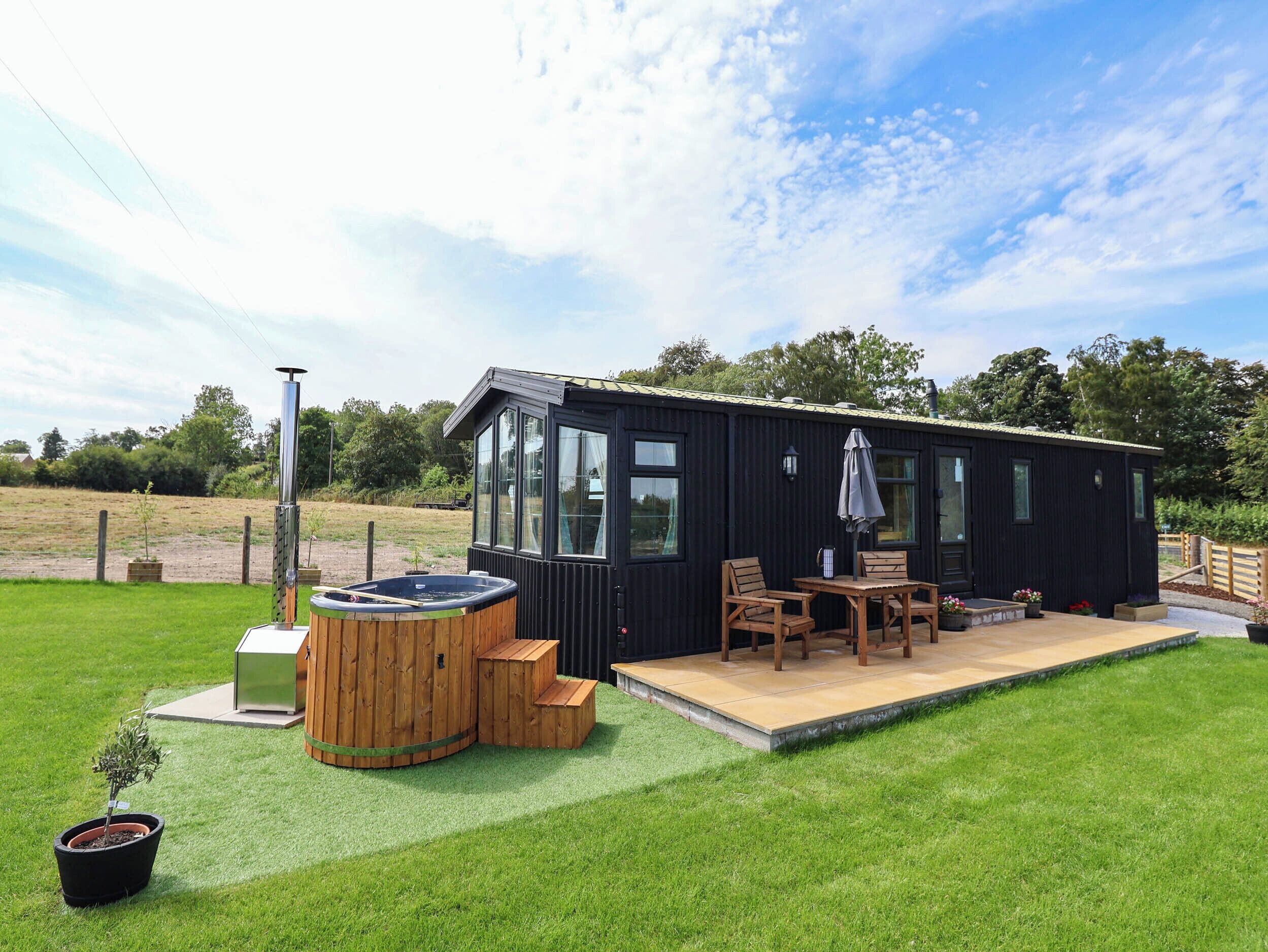A black cabin with a hot tub, wooden deck, and outdoor seating area.