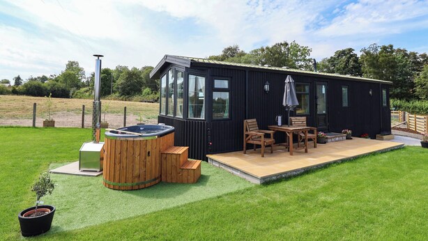 A black cabin with a hot tub, wooden deck, and outdoor seating area. - Balwen (Mold)