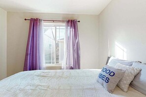 A bedroom with a bed, a window with purple curtains, and a pillow with 'GOOD VIBES' text.