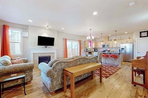 A living room with a fireplace, sofas, a wooden table, and a kitchen area with a refrigerator, microwave, and bar stools.