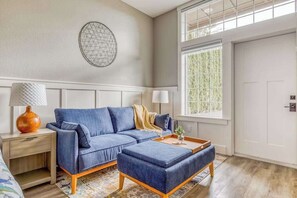 A modern living room with a blue sofa, a matching ottoman, a side table with a lamp, and a decorative wall piece. - Updated Studio Oregon Wine Country - Kitchenette (McMinnville)