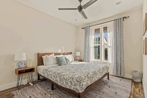 A bedroom with a bed, two bedside tables, a ceiling fan, a window with curtains, and a wooden floor.
