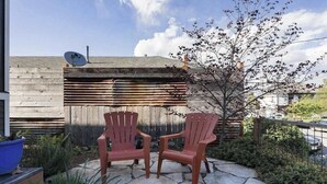 To Adirondack-stole på en stenbelagt terrasse med et træhegn og et træ i baggrunden.