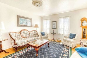 Ein Wohnzimmer mit einem blumengedeckten Sofa, einem Holztisch, zwei Sesseln, einer Uhr und einem gemusterten Teppich.