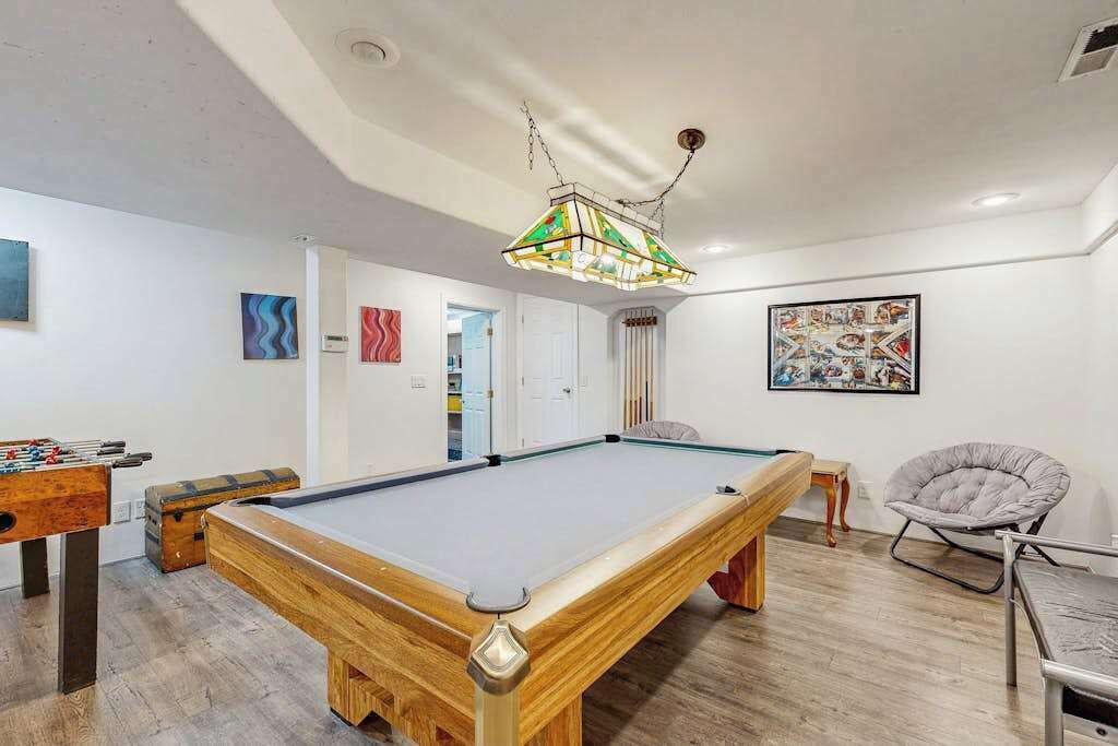Een pooltafel met houten frame, een tafelvoetbaltafel en een kleurrijke hanglamp in een kamer met witte muren en houten vloer.