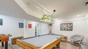 Ein Billardtisch mit Holzrahmen, ein Kicker und eine farbenfrohe Pendelleuchte in einem Raum mit weißen Wänden und Holzböden.