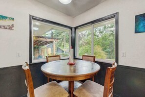 A corner dining area with a round table and four chairs, large windows with a view of greenery and a building, and a single candle on the table.