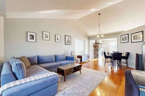 A spacious living room with a blue sectional sofa, a wooden coffee table, and a dining area with a chandelier. - 3BR Oregon Wine Country - Fireplace - Patio (McMinnville)