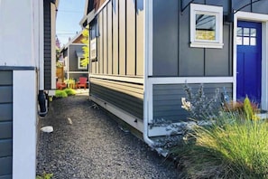 A narrow pathway leading to a house with a blue door, surrounded by gravel and some vegetation. - Micro-Loft Surrounded by Gardens in Hip Location (Portland)