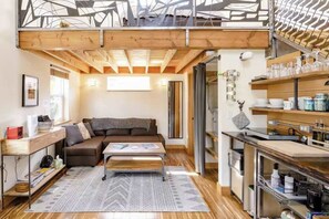 A loft-style living space with a high ceiling, a sofa, a coffee table, and a kitchen area with shelves and appliances. - Micro-Loft Surrounded by Gardens in Hip Location (Portland)
