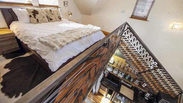 A bedroom with a bed, a wooden headboard, a nightstand, and a view of the kitchen area. - Micro-Loft Surrounded by Gardens in Hip Location (Portland)