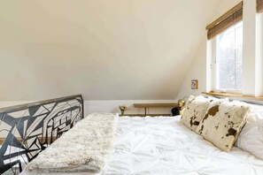 A bedroom with a bed, a fluffy white throw, a geometric headboard, a window with blinds, and a bedside table with a lamp. - Micro-Loft Surrounded by Gardens in Hip Location (Portland)
