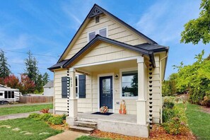 A single-story house with a front porch, a dark-colored door, and a small garden area with plants and a decorative item. - 3 BR - Downtown Newberg - Wine Tasting - WiFi (Newberg)