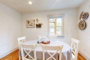 A dining area with a round table, white chairs, a window with a view, and wall decorations including a bicycle and plates. - 3 BR - Downtown Newberg - Wine Tasting - WiFi (Newberg)