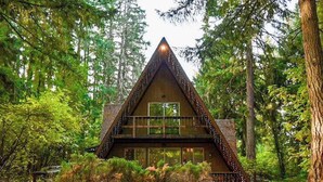 A cabin with a triangular roof, a balcony, and large windows, surrounded by dense forest. - 2BR cabin with loft in wooded setting with WiFi (Beaverton)