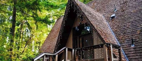 Una cabaña de madera con techo inclinado, un porche con barandilla y una ventana adornada con luces colgantes.