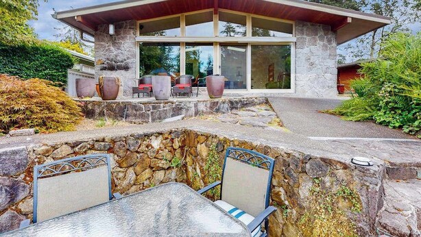 Ein modernes Haus mit Natursteinaußenwand, einer überdachten Terrasse mit Gartenmöbeln und einer Steinmauer zur Geländeanhebung.