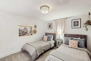 Et soveværelse med to senge, et natbord, en lampe samt vægdekorationer, heriblandt et billede i ramme og et gobelin.