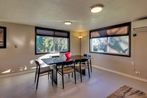 Een eethoek met een tafel, stoelen, grote ramen met zonwering en een vloerlamp.
