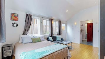 A bedroom with a bed, bedside table, and a fan. There are windows with curtains, a door, and a hallway leading to another room.
