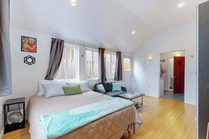 Een slaapkamer met een bed, nachtkastje en ventilator. Er zijn ramen met gordijnen, een deur en een gang die naar een andere kamer leidt.