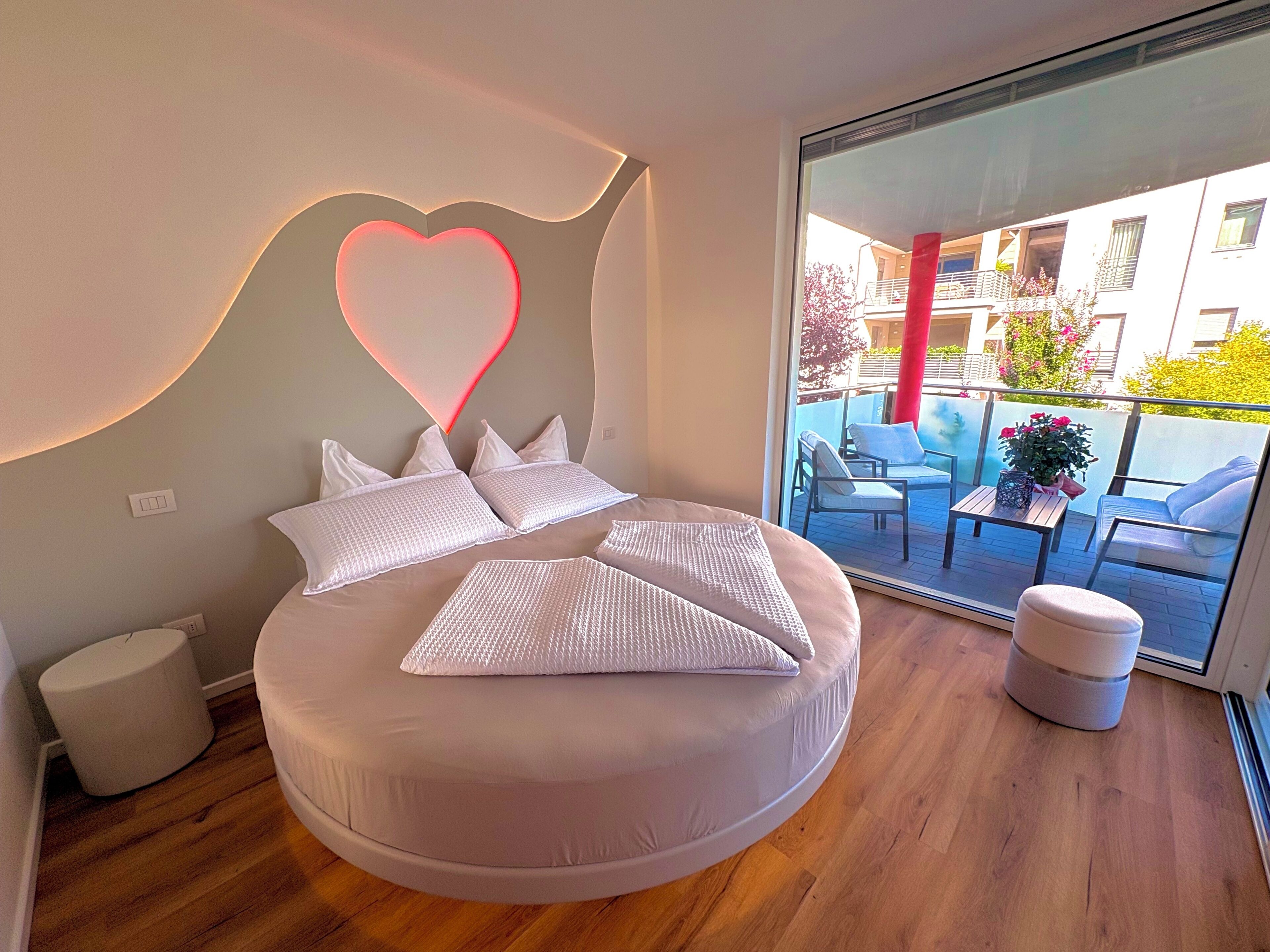 Une chambre moderne avec un lit rond, un luminaire en forme de cœur et une vue sur un balcon extérieur.