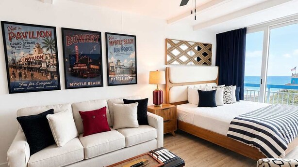 Ein Hotelzimmer mit einem Bett, einem Sofa, einem Couchtisch, einem Beistelltisch und einem Deckenventilator. An der Wand hängen drei Poster.