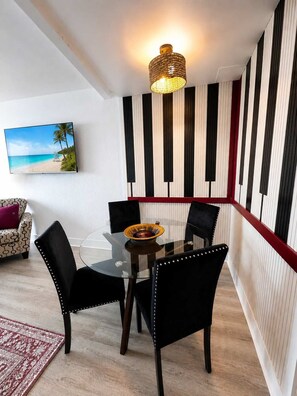 A dining area with a glass table, black chairs, a woven light fixture, and a striped wall with a beach scene painting. - Boardwalk Noir - Oceanfront Studio Yachtsman 404 (Myrtle Beach)