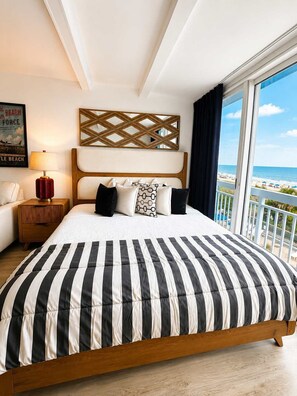 A hotel room with a large bed, a view of the beach, a wooden headboard, and a nightstand with a lamp. - Boardwalk Noir - Oceanfront Studio Yachtsman 404 (Myrtle Beach)