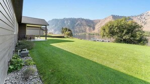 Ein gepflegter Rasen mit einer Terrassenfläche, einem Gewässer und Hügeln im Hintergrund.
