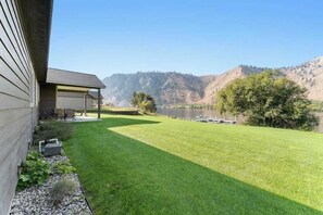 Ein gepflegter Rasen mit einer Terrassenfläche, einem Gewässer und Hügeln im Hintergrund.