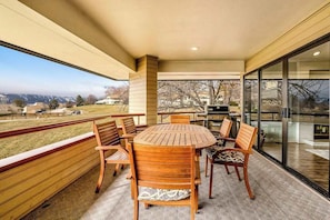 A covered outdoor patio with a round wooden table and chairs, a glass door leading to a kitchen, and a view of a residential area. - Renovated 3BR Mountainview - Pool - Hot Tub (Manson)