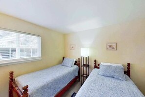Quarto com duas camas de solteiro, janela com persianas e uma mesa pequena com abajur.