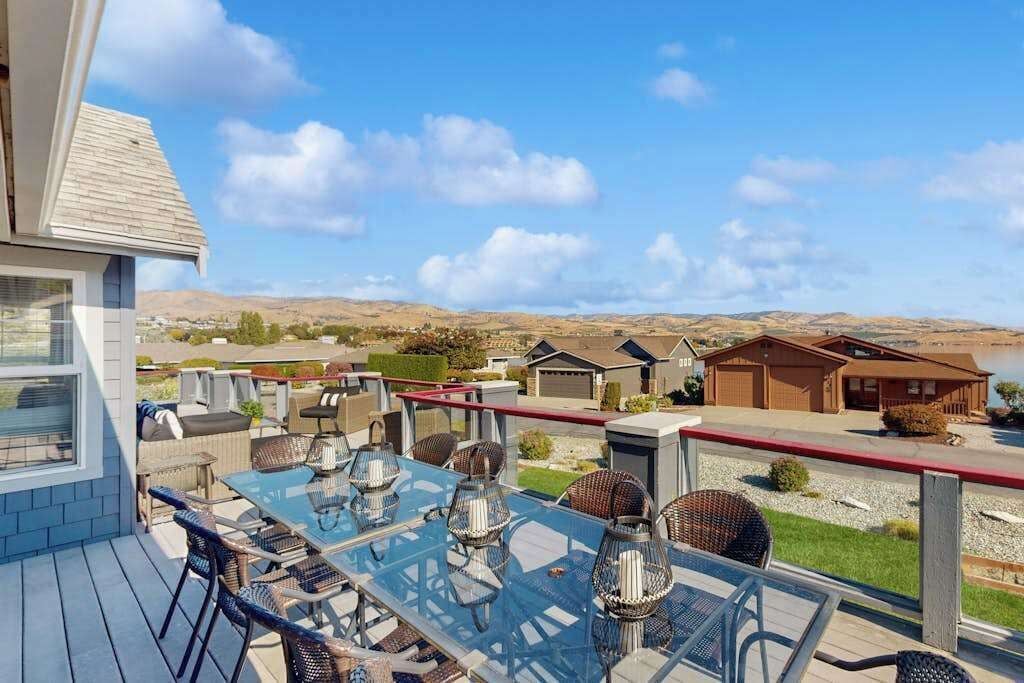 A deck with a glass table and chairs, overlooking a residential area with houses and hills.