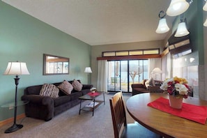 Ein Wohnzimmer mit Sofa, Couchtisch und Esstisch mit Stühlen. Es gibt ein großes Fenster mit Vorhängen, eine Lampe und ein gerahmtes Bild an der Wand.