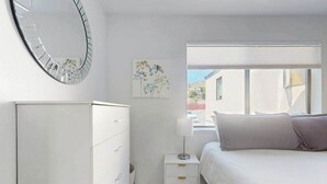 Ein Schlafzimmer mit einem Bett, einem Nachttisch mit Lampe, einer Kommode, einer Wanduhr und einem Fenster mit Jalousien.