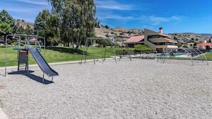 Ein Spielplatz mit Schaukeln und einer Rutsche, umgeben von Kies und Bäumen, mit Häusern im Hintergrund.