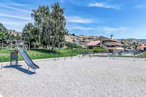 Ein Spielplatz mit Rutsche und Schaukeln, umgeben von einer Rasenfläche und einem Gebäude mit rotem Dach.