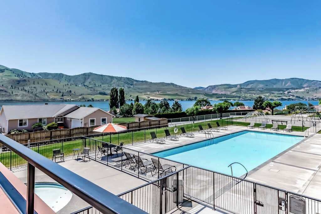 A swimming pool with a view of hills and a lake, surrounded by a fenced patio area with lounge chairs.